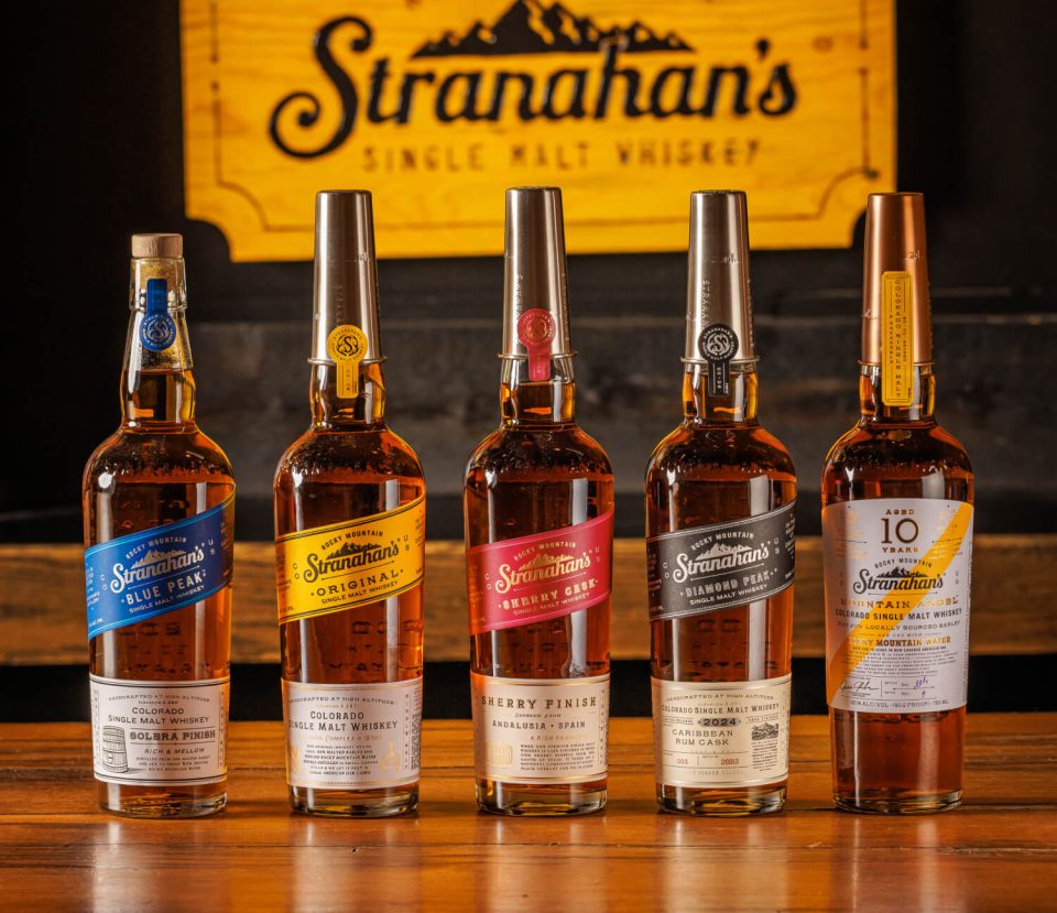 A row of a variety of Stranahan's Whiskey flavors displayed in front of a brand sign.