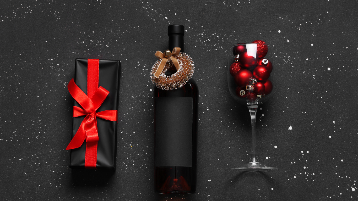 Stylish holiday-themed flat lay featuring a wrapped wine gift box with red ribbon, a wine bottle adorned with a festive wreath, and a wine glass filled with red Christmas ornaments on a black background with white snow-like speckles—perfect for seasonal gift inspiration.